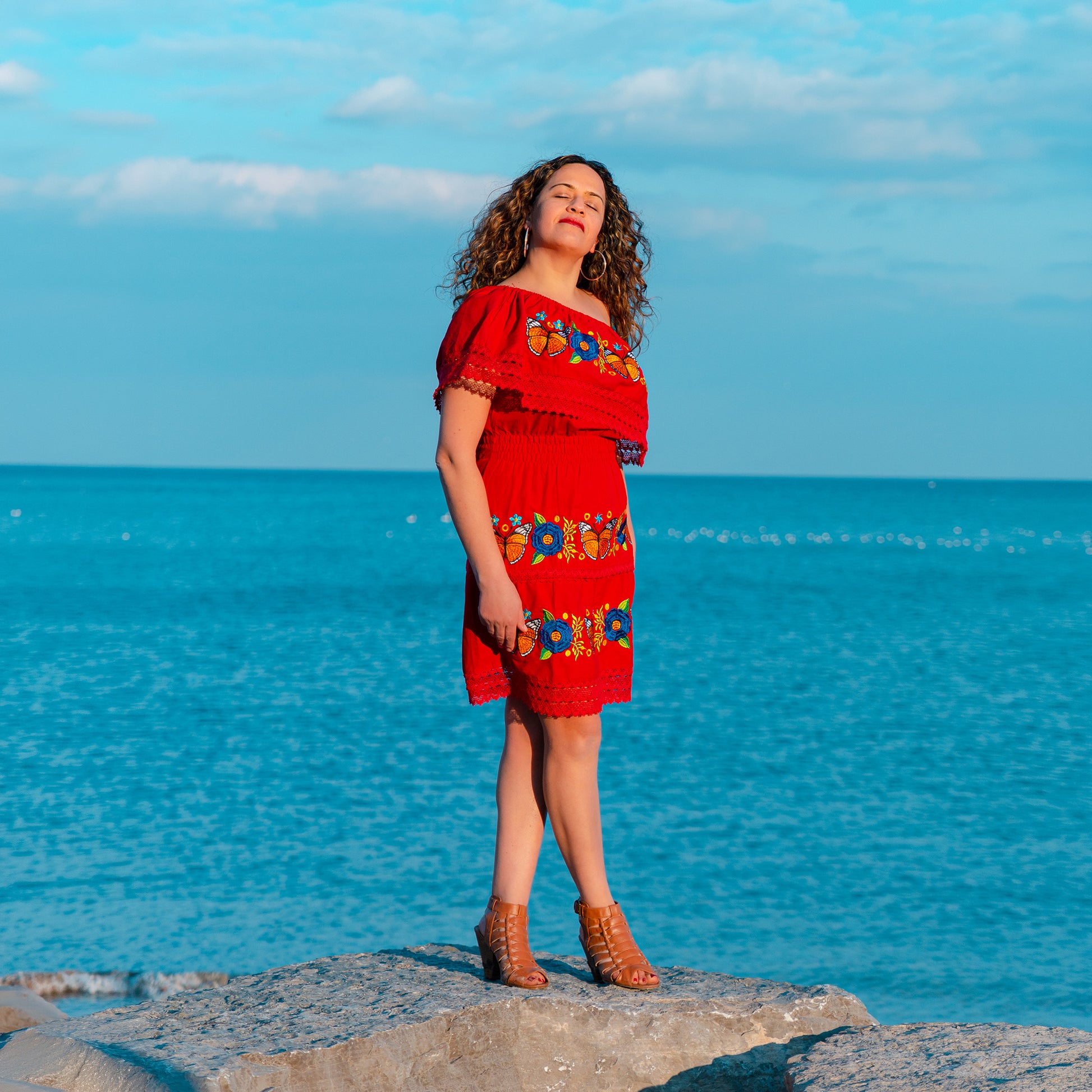 Woman in a red dress with floral patterns standing on rocks by the ocean under a blue sky.