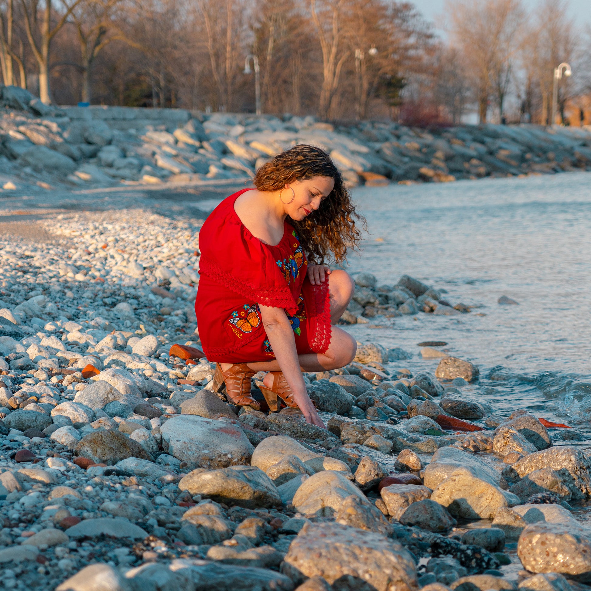 Woman wearing embroidered butterfly red dress with floral design near water at sunset
